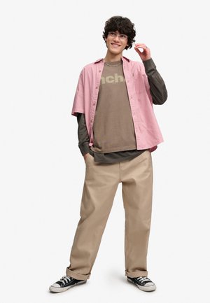 Ung person med krøllet hår iført briller, lag-på-lag afslappet tøj i pink, brun og beige, sorte sneakers, stående med den ene hånd i lommen.