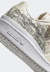 Cream-colored sneaker with black abstract line designs, textured white sole, and prominent branding. Smooth material with a modern shape.