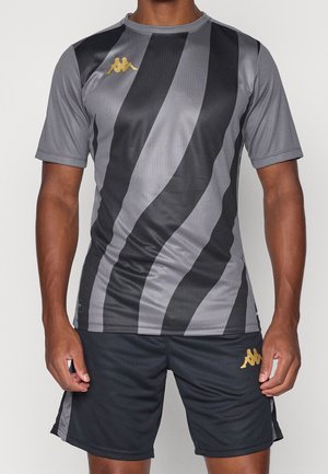 Mand iført grå og sort stribet sportstrøje og sorte shorts med gyldne Kappa-logoer, stående med armene afslappet ned langs siderne.