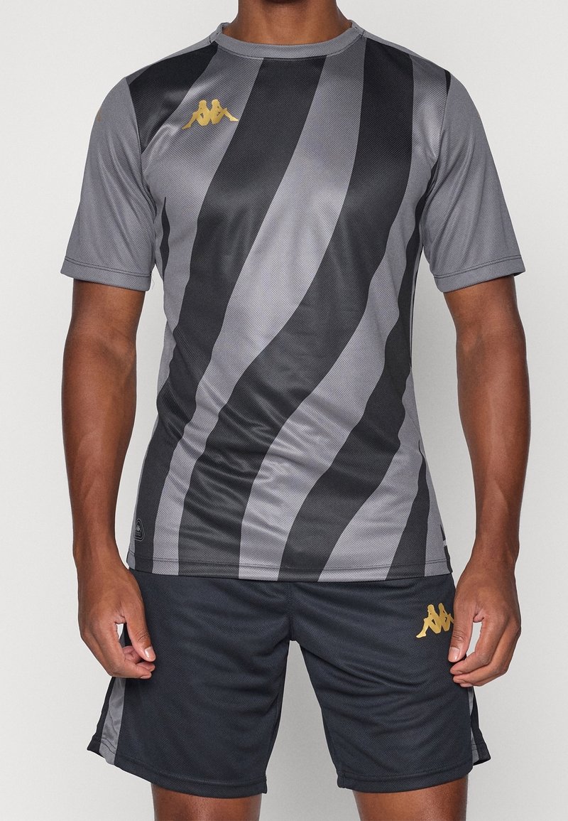 Homme portant un t-shirt de sport rayé gris et noir et un short noir avec des logos Kappa dorés, debout les bras détendus le long du corps.
