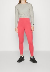Person i grå långärmad kort topp, högmidjade korall leggings med Champion-logotyper, vita strumpor och svarta träningsskor som står på grått golv.