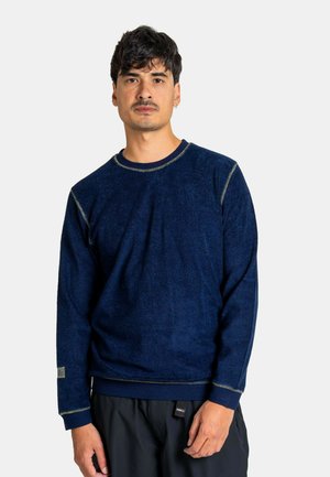 Mann mit kurzen dunklen Haaren, der ein marineblaues Langarmshirt mit Rundhalsausschnitt und schwarze Hosen trägt, steht vor einem weißen Hintergrund.