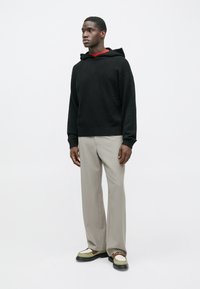 Schwarzer Strick-Hoodie mit lockerem Schnitt, gerippten Bündchen, getragen über einem roten Hemd. Helle, graue, weite Hosen und mehrfarbige Schuhe vervollständigen das Outfit.