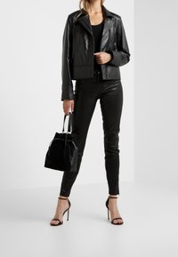 Kobieta ubrana w czarną skórzaną kurtkę, czarne dopasowane spodnie, trzymająca czarną torebkę oraz nosząca czarne sandały na obcasie, stoi w białym pomieszczeniu.