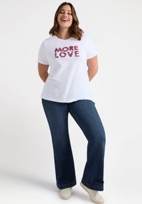 Maglietta bianca con testo floreale "MORE LOVE", maniche corte, abbinata a jeans svasati blu scuro e sneaker bianche.