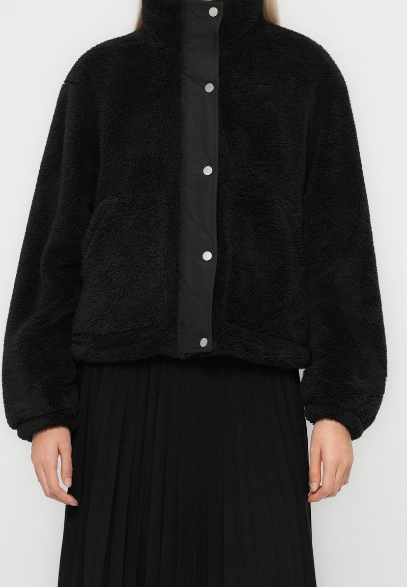 Veste polaire noire avec boutons-pression et grandes poches avant portée sur une jupe plissée noire, vue des épaules jusqu'aux hanches.