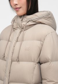Beige puffer jacket with a hood, featuring stitched detailing, drawstring accents, and a subtle logo print on the chest. Smooth texture.