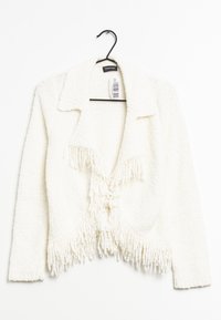 Cardigan bianco corto realizzato in un tessuto testurizzato e morbido. Presenta un colletto, frange sull'orlo e una chiusura con lacci frontali. Appeso a un gancio nero.