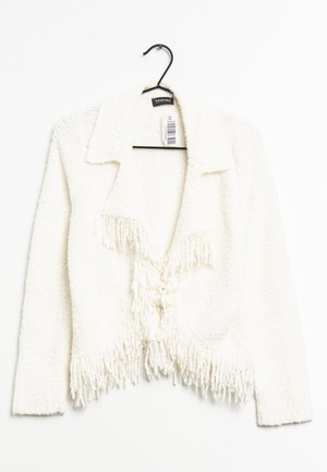 Cardigan blanc court avec un tissu texturé et duveteux. Il a un col, des franges au bas et une fermeture à nouer à l'avant. Accroché à un crochet noir.