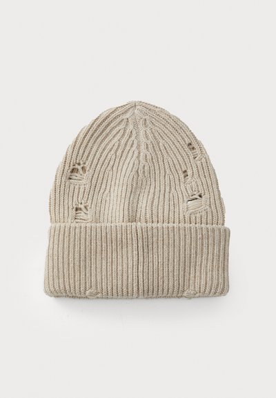 Bonnet en tricot beige avec texture côtelée, revers retourné et petits trous usés sur les côtés. Matériau doux, forme cylindrique.