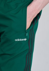 Žalios Adidas kelnės su užtrauktukų kišenėmis, su baltu logotipu ir juodomis šoninėmis juostelėmis, pagamintos iš lygaus, elastingo audinio.