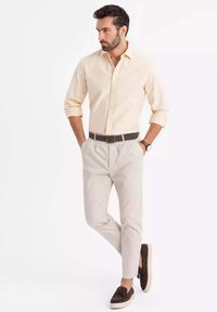 Helle beige Knopf-down-Hemd, cremefarbene Hose, brauner Gürtel und dunkle Loafers. Schlichte Gestaltung mit hochgeschobenen Ärmeln und einer Uhr am Handgelenk.