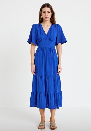 Kvinde iført en blå lagdelt midi-kjole med flagrende ærmer, V-hals, guldringøreringe, armbånd og sandaler med remme, stående foran en hvid baggrund.