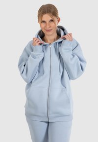 Felpa con zip di colore azzurro chiaro in tessuto morbido, dotata di un ampio cappuccio, tasche a marsupio e polsini a costine, indossata da una persona che sta aggiustando il cappuccio.