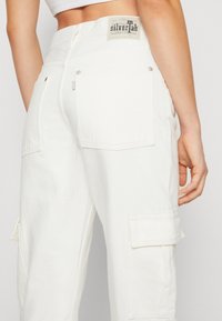 Vita cargobyxor i denim med en bakficka, silverdetaljer och en texturerad etikett vid midjan med varumärkesinformation.
