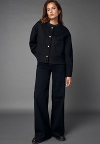 Schwarze Wollmischung Jacke mit Rundhalsausschnitt, Knopfverschluss und zwei Vordertaschen. Kombiniert mit weiten schwarzen Hosen und glänzenden Schuhen.