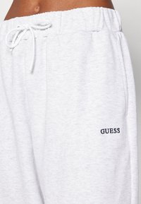 Ljusgrå sweatpants i bomullsblandning med elastisk midja och snörning samt en mörk broderad "GUESS"-logotyp på vänster sida.