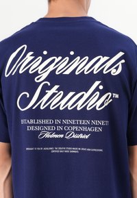 Tamsiai mėlynas medvilninis marškinėliai su baltais užrašais „Originals Studio“ ir „Įkurtas 1990 metais“ ant nugaros. Laisvas siluetas.