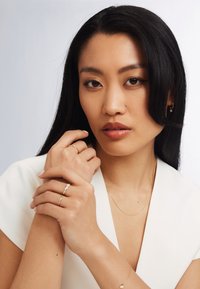 Junge Frau mit langen schwarzen Haaren, gekleidet in Weiß, zeigt goldene Ringe, ein Armband, eine Halskette und kleine Creolen vor einem schlichten Hintergrund.