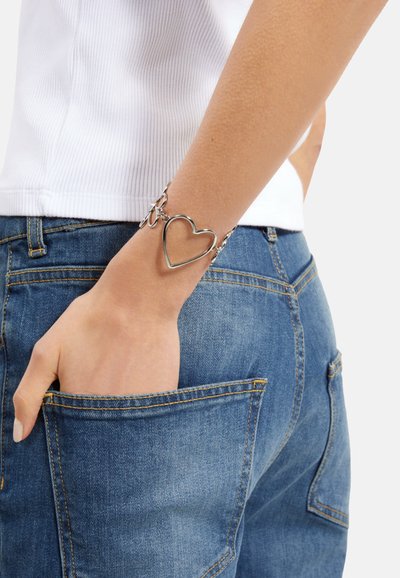 Pulsera en tono plateado con un charm en forma de corazón, cadenas entrelazadas, llevada en la muñeca. Jeans de mezclilla azules con bolsillos visibles en el fondo.