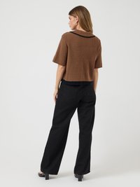 Mulher de costas, vestindo uma blusa de malha curta castanha com acabamento preto, calças largas pretas e sapatos de salto pretos, em um fundo branco.