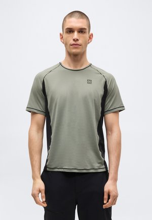 Homme debout face à l'avant, portant un t-shirt de sport gris et noir à manches courtes et un pantalon noir sur un fond blanc uni.