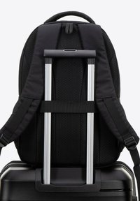 Mochila negra con textura de malla, correas ajustables y asa retráctil, apoyada sobre una maleta rígida negra. Visibles acentos de hardware plateado.