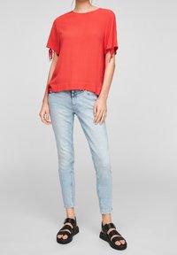 Top rojo, holgado, con mangas cortas y lazos en los laterales, combinado con jeans azul claro. Sandalias negras de tiras completan el conjunto.
