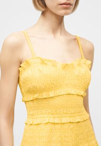 Γυναίκα που φοράει κίτρινο αμάνικο φόρεμα με βολάν στις άκρες και λεπτομέρειες από ρυτιδιασμένο ύφασμα, κομμένο κάτω από το πηγούνι και πάνω από τη μέση.