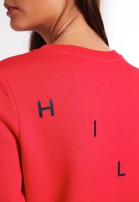 Červená mikina s žebrovaným výstřihem a grafickým motivem s černými písmeny "H", "I" a "L" na zádech. Hladká textura.