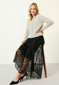 Hellgraue Strickjacke mit rundem Halsausschnitt und Knöpfen, kombiniert mit einem schwarzen Spitz Rock mit komplexen Mustern und gefranstem Saum. Schwarze Absätze.