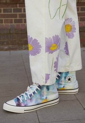 Hvide bukser med store lilla og gule blomster kombineret med blå og hvide tie-dye høje Converse-sneakers på grå asfalt.