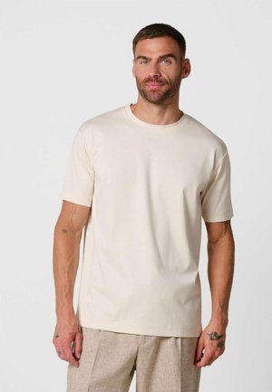 Mann mit kurzen Haaren und Bart, der ein schlichtes beige Kurzarm-T-Shirt und hellbraune Hose vor neutralem Hintergrund trägt.