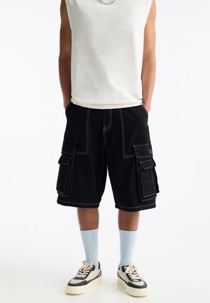 Person mit ärmellosem weißen Shirt, schwarzen Cargoshorts mit weißen Nähten, hellblauen Socken und weißen Sneakers, Hände in den Taschen.