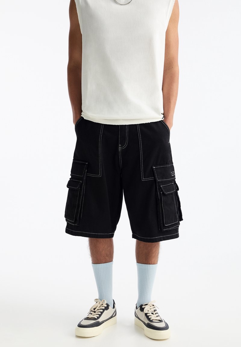Personne portant un débardeur blanc, un short cargo noir avec des coutures blanches, des chaussettes bleu clair et des baskets blanches, les mains dans les poches.