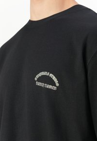 Sort crewneck t-shirt med hvid broderet tekst "Organic Studio" buet over "Daily Basics" på venstre bryst.