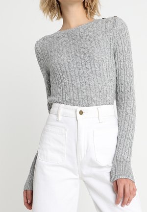 Woman wears a fitted gray cable-knit sweater with button details on shoulder and high-waisted white pants with large front pockets.