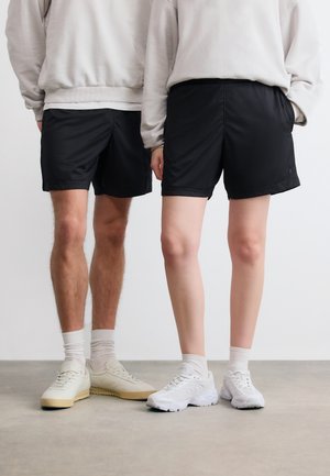 To personer står side om side iført beige sweatshirts, sorte shorts, hvide sokker og hvide sneakers på en grå gulv.