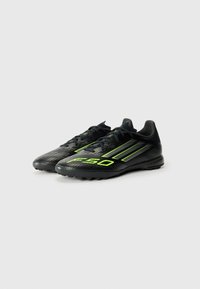 Des crampons de football noirs avec des accents verts, présentant une surface texturée, un design profil bas, un bout arrondi et des crampons en caoutchouc pour l'adhérence.