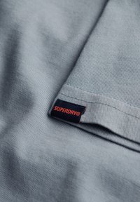 Szary prążkowany materiał bawełniany t-shirtu z granatową metką, na której widnieje napis "SUPERDRY" w kolorze pomarańczowym. Tkanina ma miękką teksturę i delikatnie się układa.