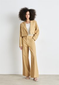 Lühendatud beež jakk ühe nööbiga, koos kõrge vöökoha ja laia jala beežide pükstega, millel on ees seam detailing ja küljetaskud.