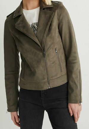 Mujer con chaqueta de cuero estilo motero en color verde oliva con bolsillos con cremallera y vaqueros negros sobre fondo neutro.