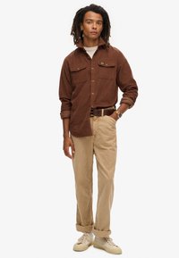 Bruine corduroy shirt met twee borstzakken, beige corduroy broek en crèmewitte sneakers. Eenvoudig horloge om de pols, staande pose.