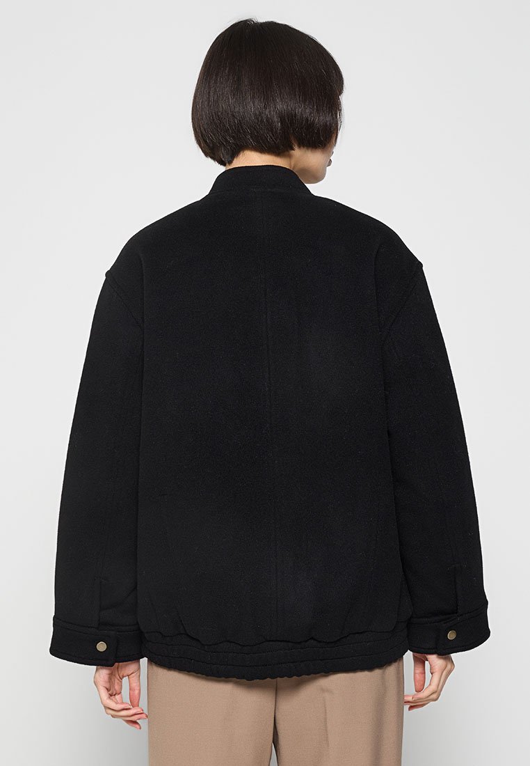 Schwarze Fleecejacke mit Stehkragen, loser Silhouette, seitlichen Taschen und Knopf-Details. Glatte Textur und minimalistisches Design.