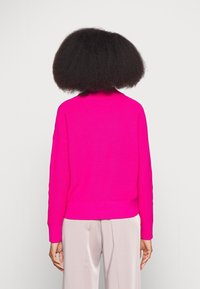 Pull en tricot rose vif à col haut, texture côtelée et manches longues, associé à un pantalon beige clair. Vue de dos présentée.