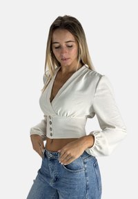 Weißes, kurzes Top mit V-Ausschnitt, Knopfdetails auf der Vorderseite und Puffärmeln, kombiniert mit hochgeschnittenen blauen Jeans. Glatte Stoffstruktur.