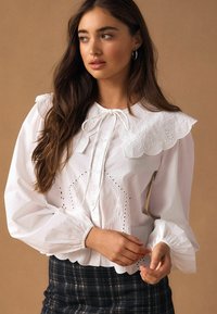 Valge pluus tikitud lainelise kraega, puffsifoonidega ja silmkoe detailidega. Omab sõlmitavat kaelust ja lühendatud, lööklaotusega äärega.