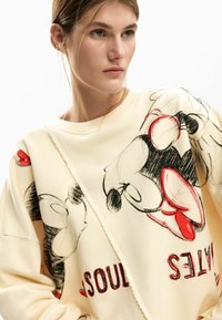 Licht crèmekleurige sweatshirt met zwart- en roodkleurige illustraties van cartoonfiguren. De stof lijkt zacht en heeft een ontspannen, oversized pasvorm.