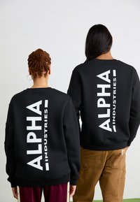 Zwarte sweaters met grote witte tekst "ALPHA INDUSTRIES" op de rug, gedragen door twee personen, één in bruine broek, eenvoudig ontwerp.