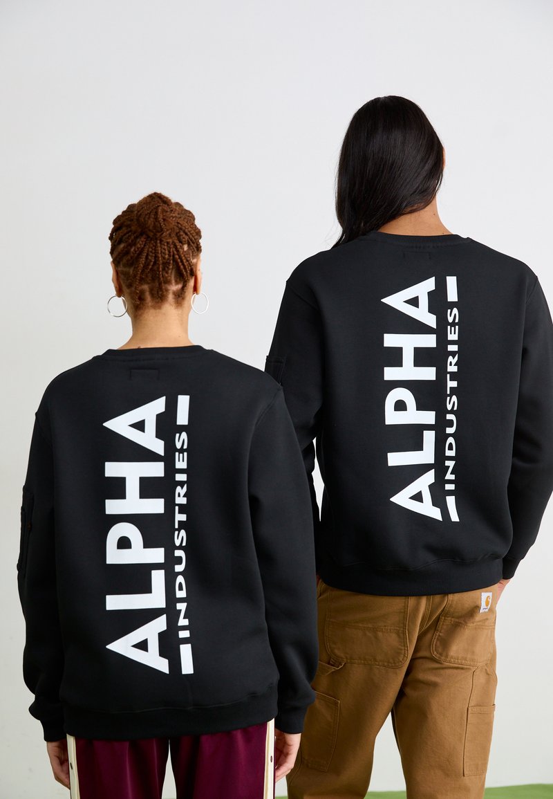 Sorte hettegensere med stor hvit "ALPHA INDUSTRIES" tekst på ryggen, båret av to personer, en i brune bukser, enkelt design.